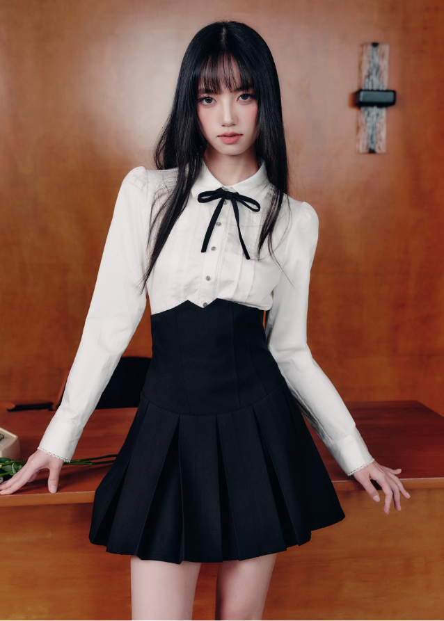 The Cherry Blossom Sonata Set captures the duality of innocence and control — a signature Fragileheart silhouette. The crisp ivory blouse, adorned with a black ribbon tie, flows into a high-waisted pleated skirt that sculpts the waist with subtle strength. Inspired by moonlit refinement and poetic order, it’s a quiet statement of authority wrapped in grace.