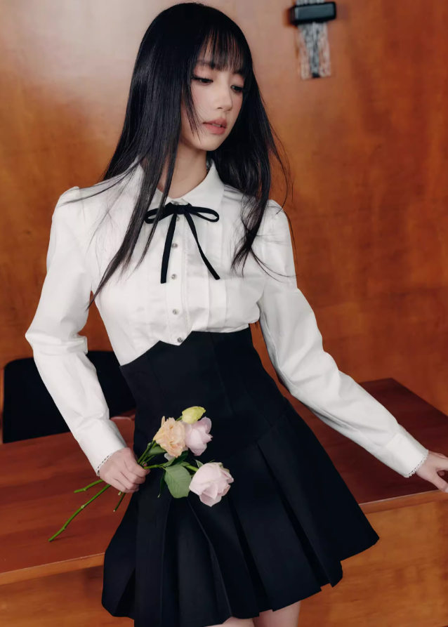 The Cherry Blossom Sonata Set captures the duality of innocence and control — a signature Fragileheart silhouette. The crisp ivory blouse, adorned with a black ribbon tie, flows into a high-waisted pleated skirt that sculpts the waist with subtle strength. Inspired by moonlit refinement and poetic order, it’s a quiet statement of authority wrapped in grace.